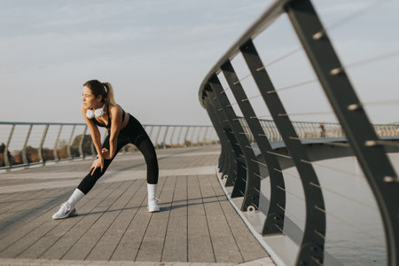 Inhaling deeply, a woman prepares her body for exercise by stretching on a sleek bridge, enjoying the tranquility of an early morning near the waterの写真素材