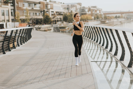 Fit woman in a black athletic outfit runs on a sleek bridge, enjoying her evening routine. The horizon glows softly, highlighting her determination and commitment to fitnessの写真素材