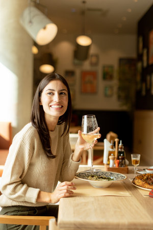 A young woman smiles brightly as she raises a glass of white wine in celebration, savoring her delicious meal in a cozy, modern restaurant filled with vibrant decor.の写真素材