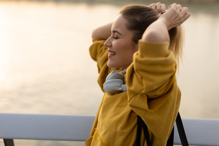 With the sun setting behind her, a cheerful young woman stands by the water, savoring a tranquil moment as she playfully pulls back her hair, feeling carefree and contentの写真素材