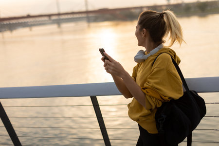As the sun sets over the tranquil river, a young woman wearing a yellow hoodie and headphones smiles while texting, capturing a moment of joy and relaxation in natureの写真素材
