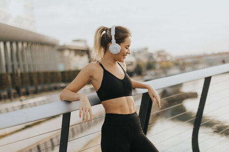 As the sun sets over the water, a young woman leans against a railing, savoring her favorite tunes through headphones, embracing tranquility and peace.の写真素材