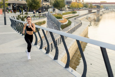 A dedicated runner maintains her pace on a riverwalk surrounded by urban architecture and lush greenery, embracing a beautiful morning and an active lifestyle.の写真素材