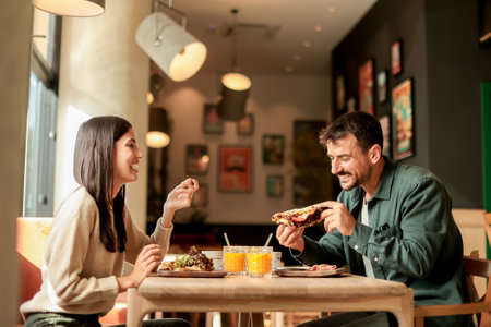 Couple shares laughter and delicious food while sitting at a wooden table in a bright cafe, surrounded by greenery and other patrons enjoying their mealsの写真素材