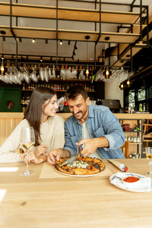 A man and woman sit at a wooden table in a welcoming restaurant, smiling as they share a tasty pizza topped with fresh ingredients, accompanied by glasses of wine.の写真素材