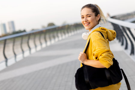 With a joyful smile, a young woman in a yellow hoodie strolls along a scenic riverside path, her black bag slung over her shoulder, embracing the gentle evening breeze.の写真素材