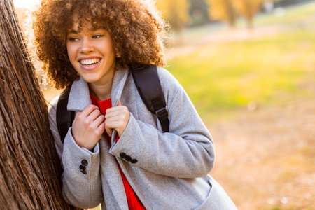 Smiling young woman with curly hair stands against a tree, wearing a cozy outfit. She embraces the beauty of autumn in a serene park, filled with colorful leaves and soft sunlight.の写真素材