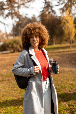 A joyful woman with curly hair strolls through a sunny park adorned with colorful autumn leaves, holding a warm cup of coffee while wearing a stylish outfit.の写真素材