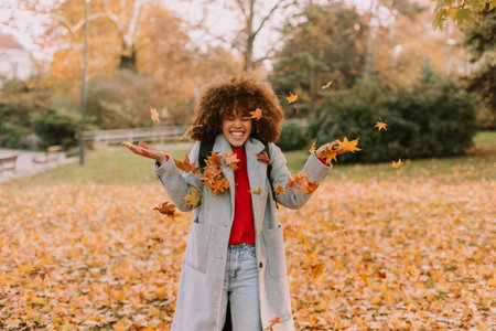 A cheerful individual with curly hair plays in a park during autumn, joyfully tossing vibrant leaves into the air among a golden blanket of fallen foliage.の写真素材