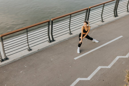 In the calm of the morning, a young woman in athletic wear stretches along a city path, her...