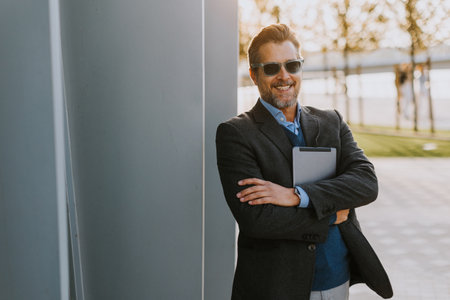 A man in business attire stands relaxed against a sleek structure, smiling while holding a tablet, embodying a blend of professionalism and leisure on a sunny day.の写真素材