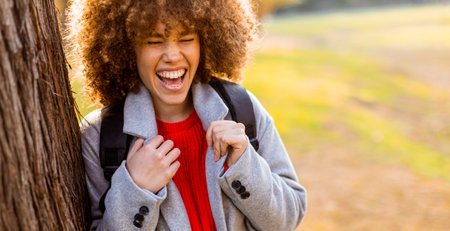 A carefree young woman laughs heartily while standing next to a tree in a serene park. The warm sunlight filters through the leaves, creating a joyful atmosphere.の写真素材