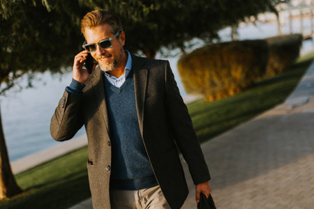 A well-dressed man strolls by the water, absorbed in a phone call. Surrounded by lush greenery, he pairs sophistication with casual ease on a sunny day.の写真素材