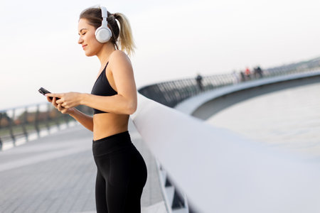 A young woman stands relaxed on a bridge, headphones on, savoring music while basking in the gentle glow of sunset near the water's edge.の写真素材