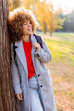 Smiling young woman with curly hair stands against a tree, wearing a cozy outfit. She embraces the beauty of autumn in a serene park, filled with colorful leaves and soft sunlight.の写真素材