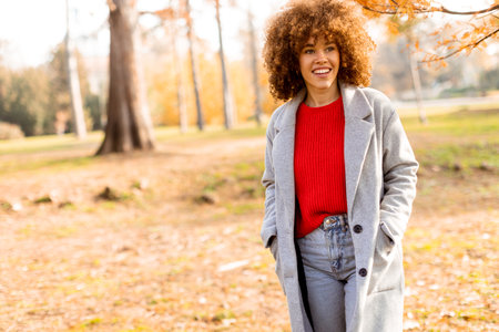 Bright autumn day captivates as a young woman walks through a lovely park, dressed in a cozy gray coat and red sweater, embodying warmth and joy amidst nature's beauty.の写真素材