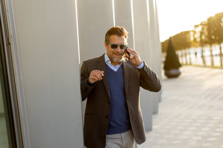 A well-dressed man enjoys a casual conversation on the phone while walking along a sunlit pathway next to contemporary buildings, creating an ambiance of sophistication.の写真素材