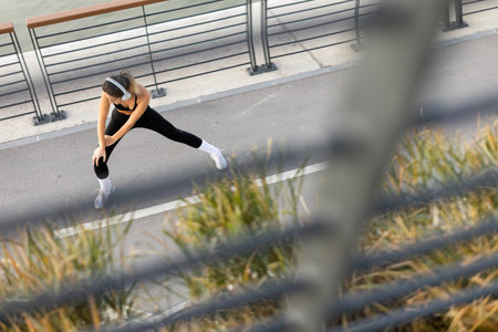 In the calm of the morning, a young woman in athletic wear stretches along a city path, her headphones on, focused on energizing her body for a workout or run.の写真素材
