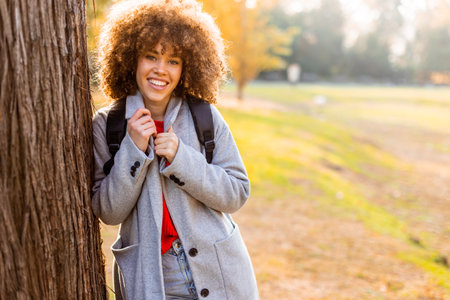 Smiling young woman with curly hair stands against a tree, wearing a cozy outfit. She embraces the beauty of autumn in a serene park, filled with colorful leaves and soft sunlight.の写真素材
