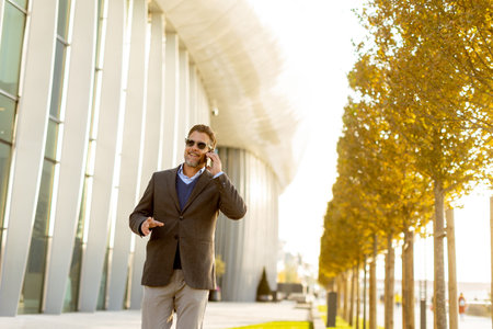 A well-dressed man strolls along a sleek pathway, chatting on his phone under the warm glow of the afternoon sun, surrounded by contemporary architecture and greenery.の写真素材