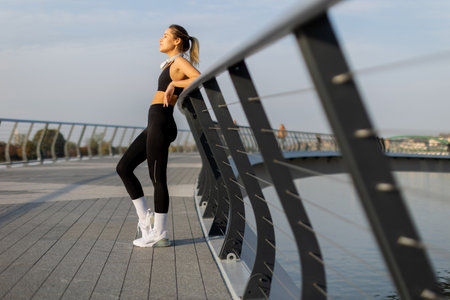 A young woman exercising on a sleek boardwalk under a clear sky, she shields her eyes from the sun, embodying a mix of focus and elegance in her activewear.の写真素材