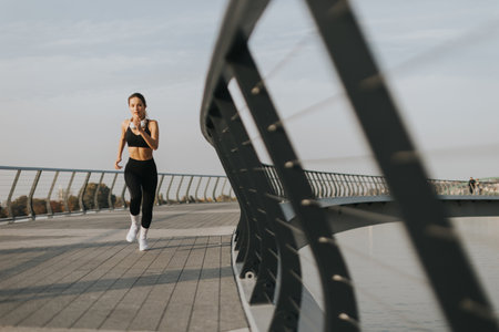 A young woman in athletic attire runs energetically along a riverside path, embracing the serene atmosphere of a beautiful morning, fueled by her favorite music.の写真素材
