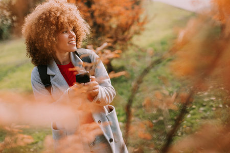 A joyful person with curly hair smiles while holding a phone, surrounded by vibrant autumn leaves in a serene outdoor setting on a sunny day.の写真素材