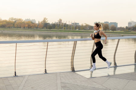 A determined runner strides confidently along a riverside path, embracing the crisp morning air, with city skyline and autumn trees providing a picturesque backdrop.の写真素材