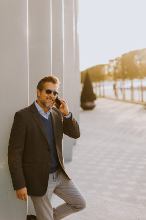 A well-dressed man is leaning casually against a sleek white wall while talking on his phone. The warm light of sunset beautifully highlights the modern architecture around him.の写真素材