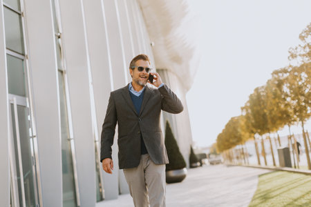 A well-dressed man strolls along a sleek pathway, chatting on his phone under the warm glow of the afternoon sun, surrounded by contemporary architecture and greenery.の写真素材