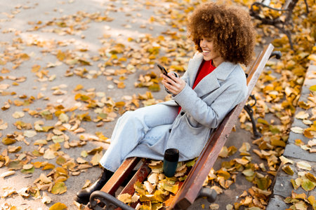 Sitting on a wooden bench in a park, a young woman smiles while texting on her smartphone, enjoying the vibrant autumn leaves that blanket the ground.の写真素材