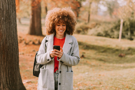 On a bright autumn day, a cheerful woman stands by a tree, absorbed in her phone while surrounded by vibrant foliage. Her cozy outfit contrasts beautifully with the seasonal colors.の写真素材