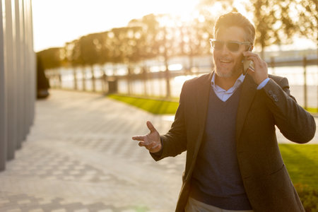 A well-dressed man enjoys a casual conversation on the phone while walking along a sunlit pathway next to contemporary buildings, creating an ambiance of sophistication.の写真素材
