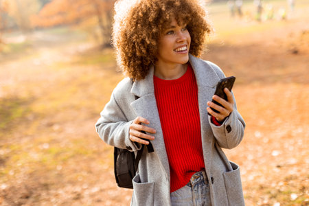 A joyful woman with curly locks is engaging with her smartphone, surrounded by the warm colors of autumn leaves in a lively outdoor park setting.の写真素材