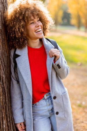 Smiling young woman with curly hair stands against a tree, wearing a cozy outfit. She embraces the beauty of autumn in a serene park, filled with colorful leaves and soft sunlight.の写真素材