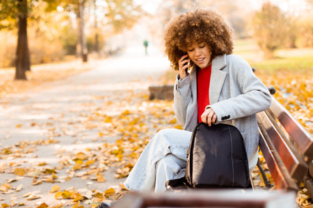 A joyful woman with curly hair makes a phone call while sitting on a park bench surrounded by vibrant autumn foliage, enjoying a beautiful day outdoors.の写真素材