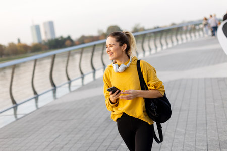 A smiling young woman walks along a riverside path in autumn, dressed in a cozy yellow sweater and headphones. The serene atmosphere captures her joyful moment.の写真素材