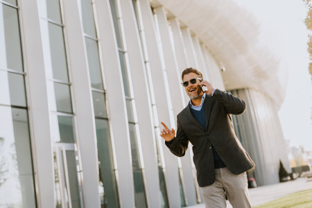 A well-dressed man strolls along a sleek pathway, chatting on his phone under the warm glow of the afternoon sun, surrounded by contemporary architecture and greenery.の写真素材