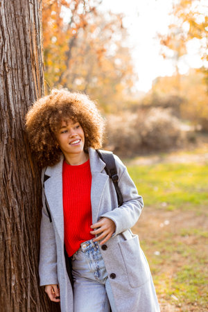Smiling young woman with curly hair stands against a tree, wearing a cozy outfit. She embraces the beauty of autumn in a serene park, filled with colorful leaves and soft sunlight.の写真素材