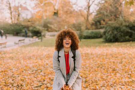 A cheerful individual with curly hair plays in a park during autumn, joyfully tossing vibrant leaves...