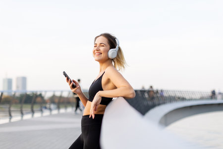 A young woman stands relaxed on a bridge, headphones on, savoring music while basking in the gentle glow of sunset near the water's edge.の写真素材