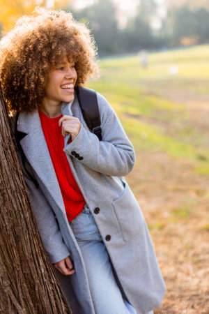 Smiling young woman with curly hair stands against a tree, wearing a cozy outfit. She embraces the beauty of autumn in a serene park, filled with colorful leaves and soft sunlight.の写真素材
