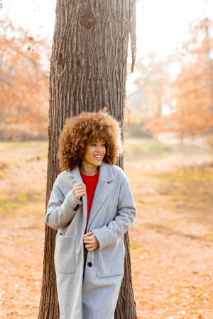 A young woman with curly hair leans against a tree, wearing a warm sweater, smiling joyfully amidst the golden autumn foliage and fallen leaves around her.の写真素材