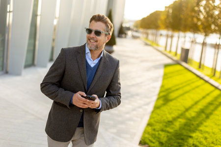 A professional man in stylish attire leisurely walks outside a sleek building, smiling as he interacts with his phone under the warm afternoon sun, exuding confidence.の写真素材