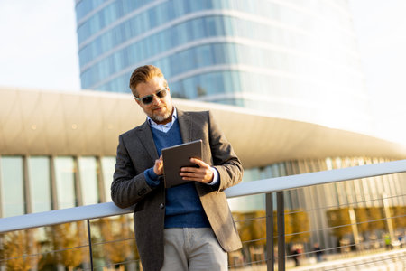 A stylish man stands on a balcony, engaging with a tablet against a backdrop of sleek architecture.の写真素材