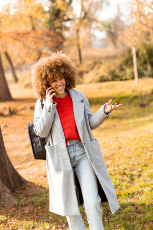 A joyful woman with curly hair dressed in a gray coat and red sweater speaks animatedly on her phone while enjoying the beautiful autumn colors in a serene park.の写真素材