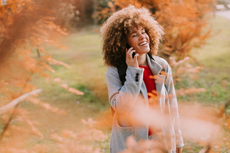 A young woman with curly hair smiles while talking on her phone, immersed in beautiful autumn colors. The autumn leaves create a warm and inviting atmosphere.の写真素材