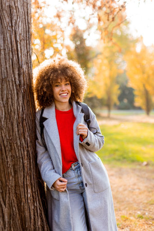 Smiling young woman with curly hair stands against a tree, wearing a cozy outfit. She embraces the beauty of autumn in a serene park, filled with colorful leaves and soft sunlight.の写真素材