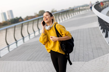 A cheerful woman dressed in a vibrant yellow sweater laughs on her phone while strolling along a picturesque riverside walkway, enjoying the warm sunshine.の写真素材