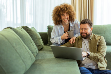 In a stylish living room, a young couple shares a joyful moment as they collaborate on a laptop, surrounded by soft green furnishings and warm sunlight streaming through the windows.の写真素材